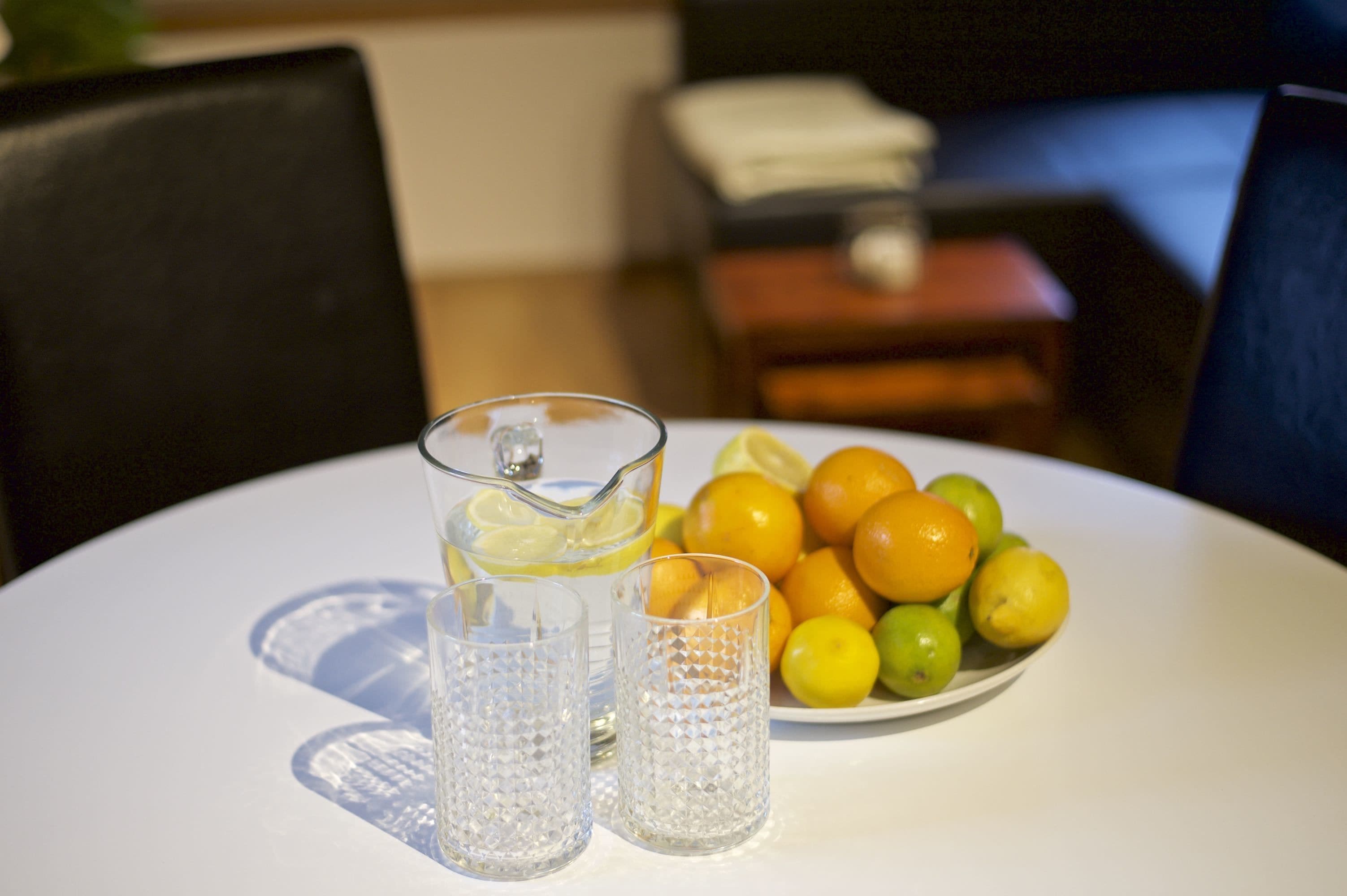 Una mesa con una jarrón de agua, dos vasos y un plato de limones y naranjas.
