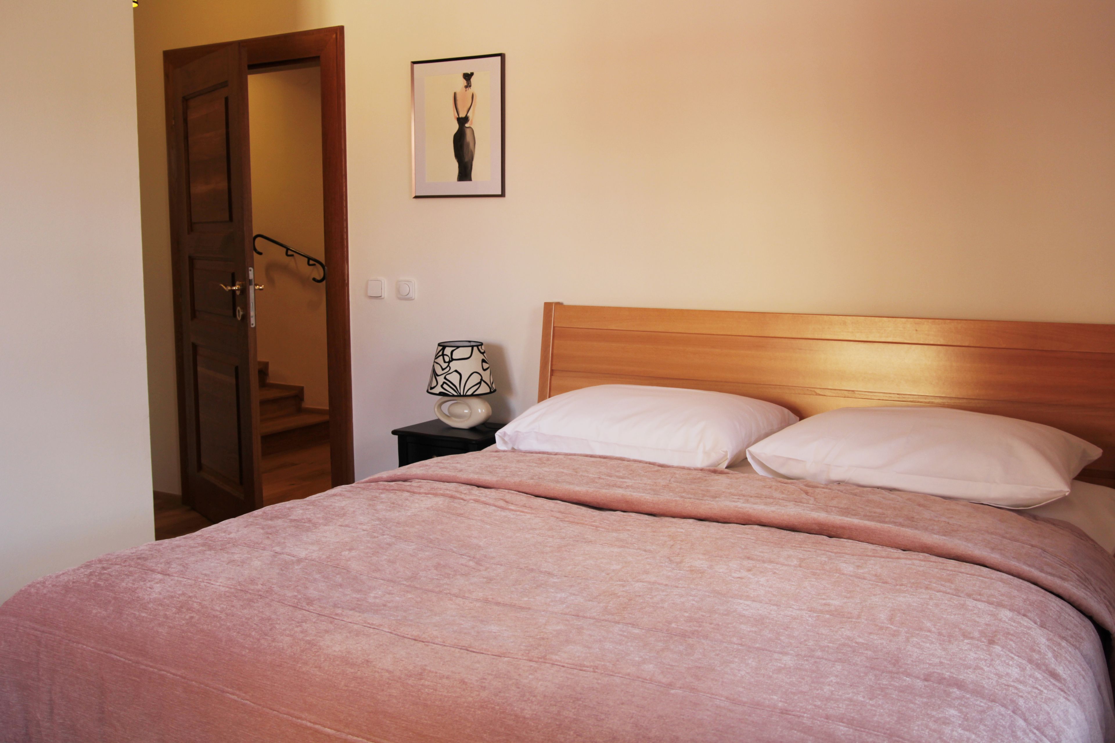 A bedroom with a wooden headboard, a bed with pink bedding, a nightstand with a lamp, and a framed picture on the wall.