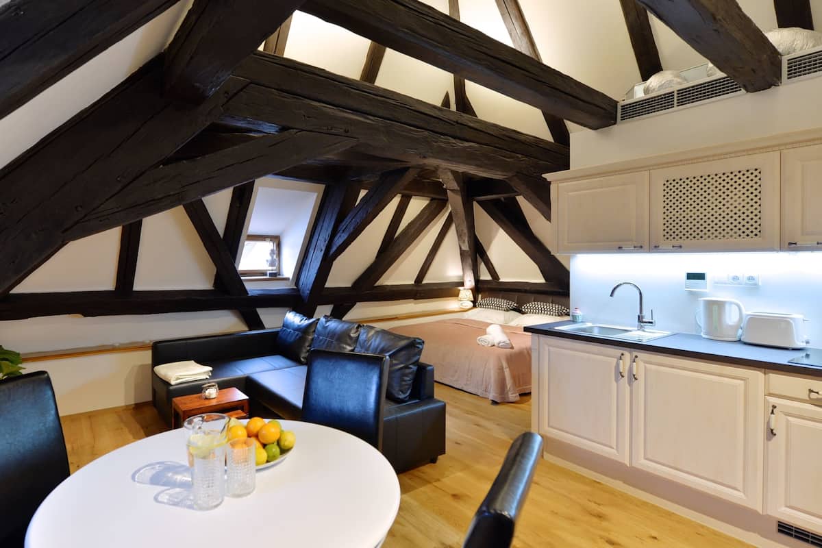 A modern loft-style room with exposed wooden beams, a cozy seating area, a kitchenette, and a view of the city.
