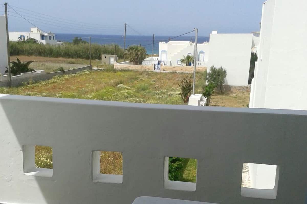 A balcony with a white table and a blue bowl, overlooking a residential area with white buildings and a view of the sea.