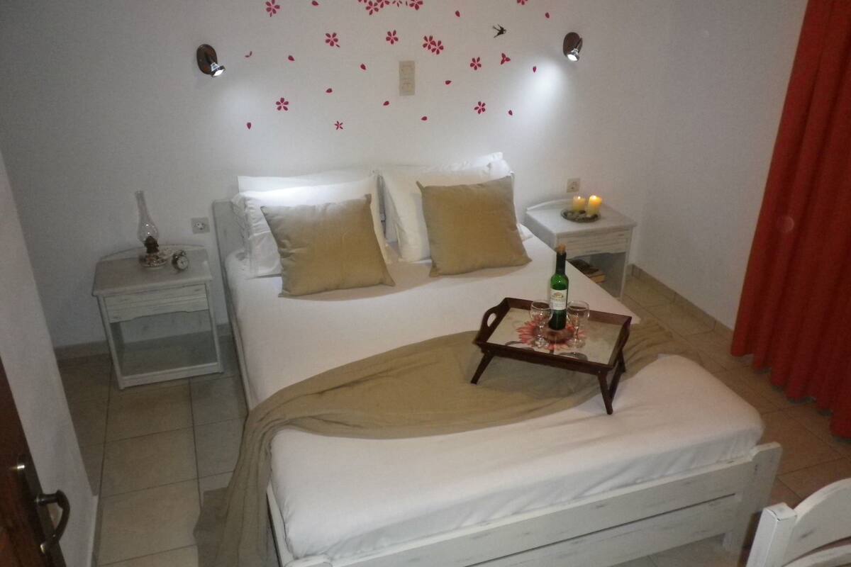 A neatly made bed with a tray of wine glasses and a bottle, a bedside table with a lamp, and a wall decorated with flower decals.