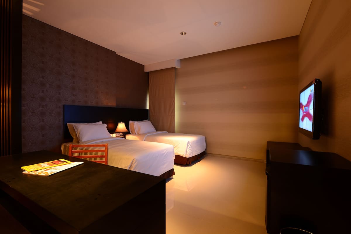A hotel room with two beds, a TV mounted on the wall, a desk with a magazine, and a lamp.