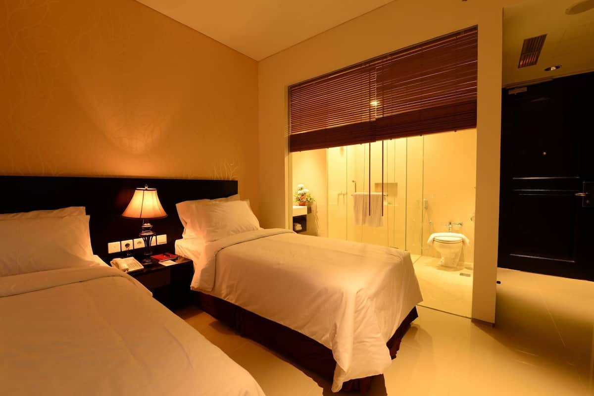 A hotel room with two beds, a nightstand with a lamp, a bathroom with a toilet, and a wardrobe.