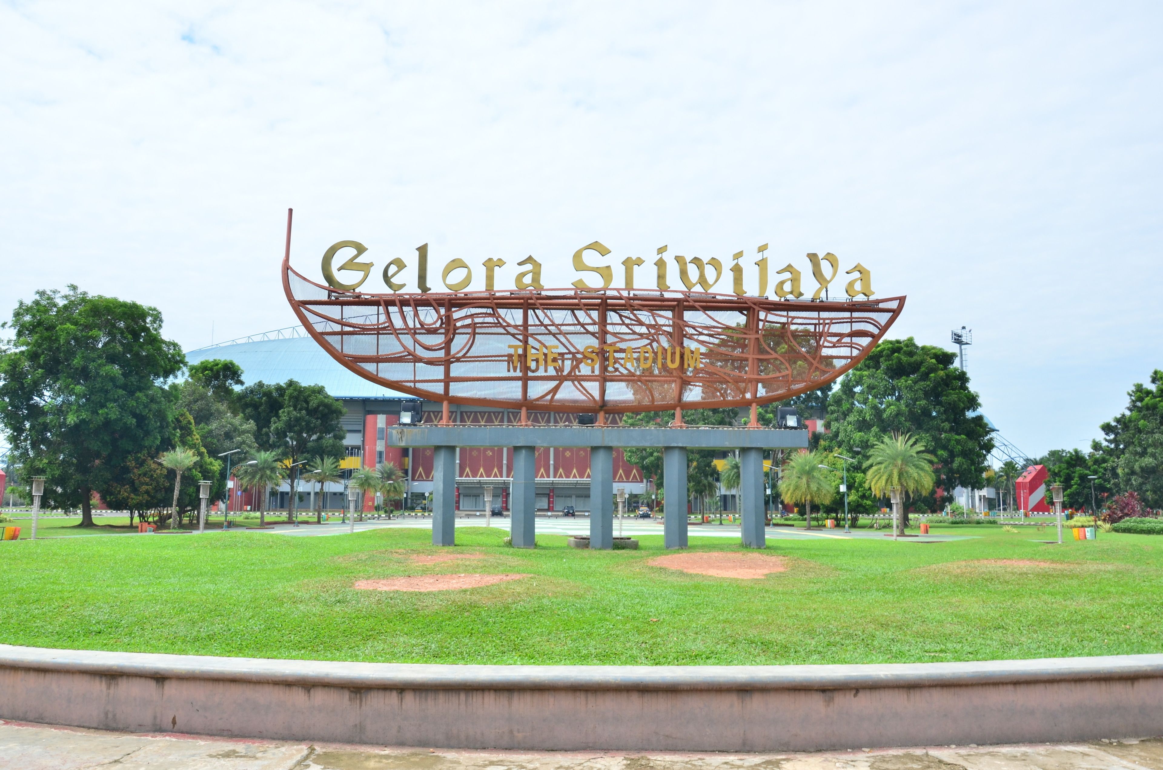 A large metal sculpture with the text "Gelora Sriwijaya Stadium" in the center, surrounded by a well-maintained grassy area and trees.