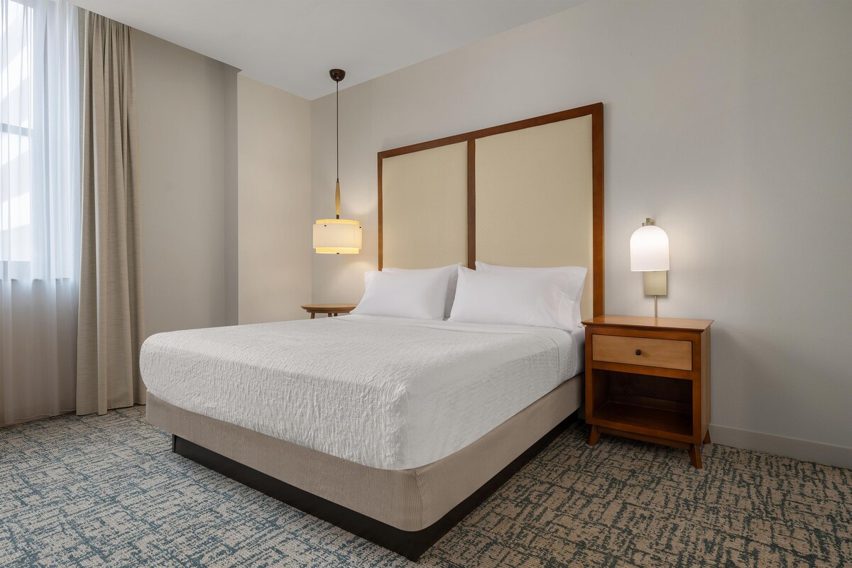 Una habitación de hotel con una cama grande, dos mesitas de noche y un ventanal con cortinas.