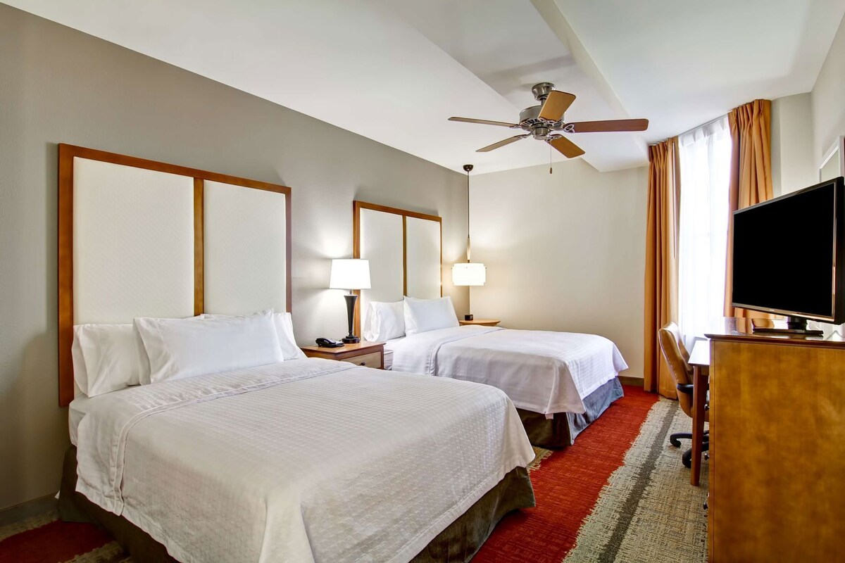 Habitación de hotel con dos camas, un escritorio con televisor, ventilador de techo y ventana con cortinas.