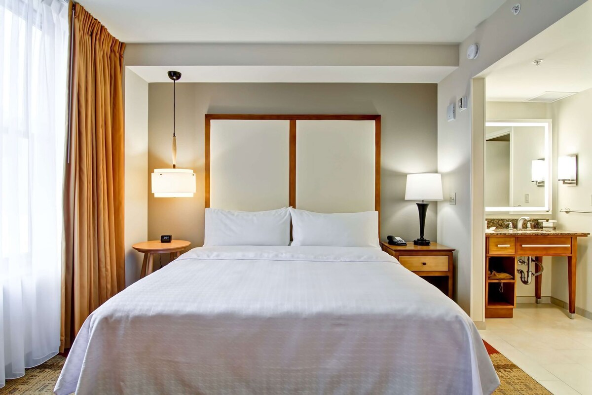Habitación de hotel con una cama grande, una mesita de noche con lámpara, un tocador con teléfono, un aparador de madera con espejo y un baño visible a través de una puerta abierta.