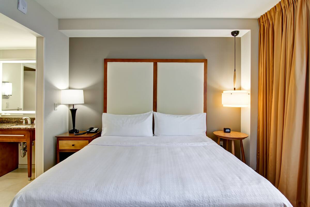 Een hotelkamer met een groot bed, een nachtkastje met een lamp, een klein tafeltje met een klok en een spiegel boven de wastafel.