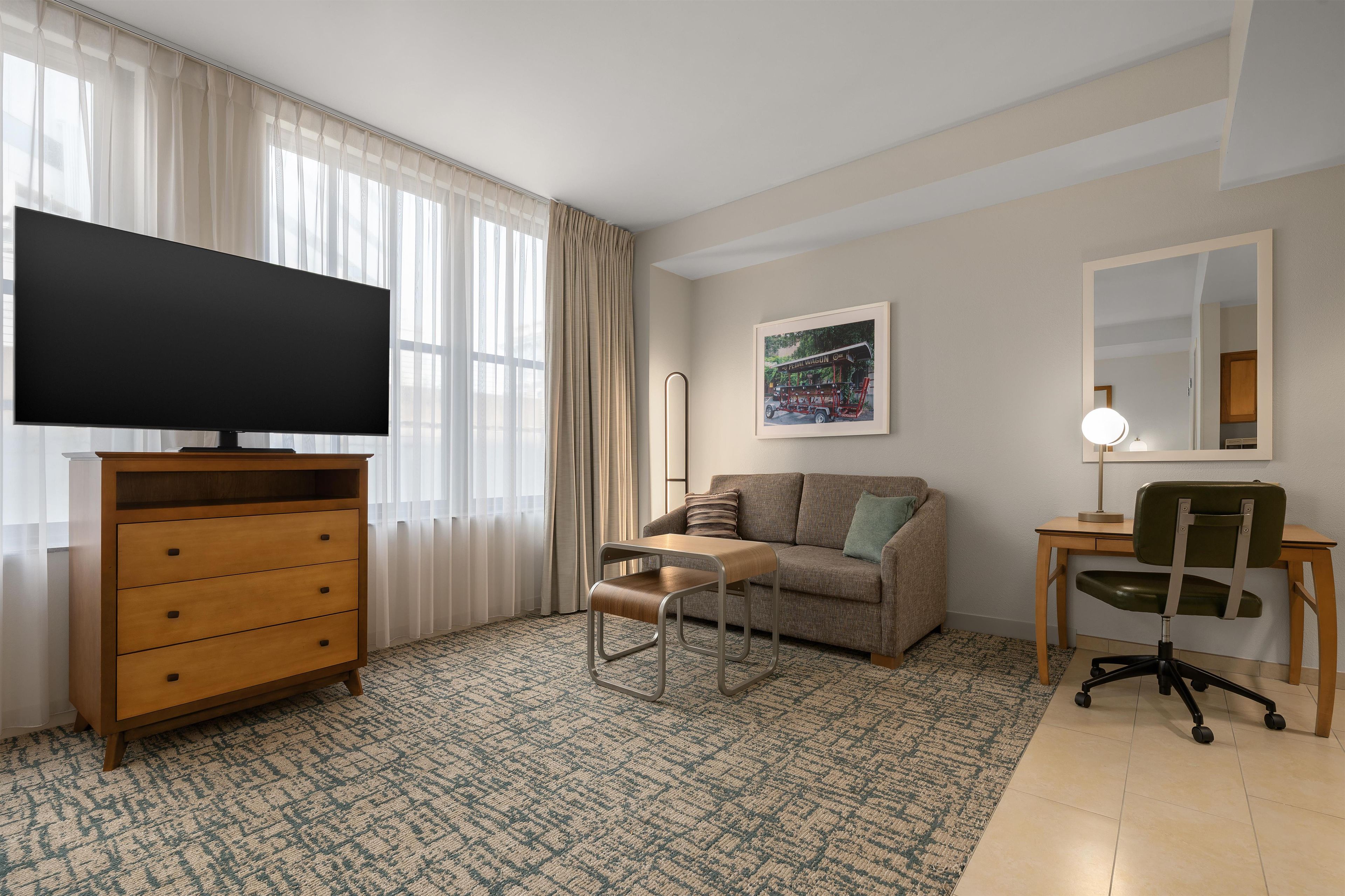A hotel room with a flat-screen TV, a wooden dresser, a sofa, a small table, a chair, and a framed picture on the wall.