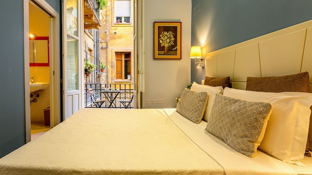 Un dormitorio con cama, almohadas, mesita de noche, lámpara de pared y puerta que da a un balcón.