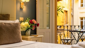 Habitación de hotel con cama, mesita de noche con un jarrón de flores, lámpara y vista al balcón exterior.