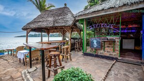 An outdoor seating area with wooden tables and chairs, a thatched-roof structure, and a view of the sea. - 2 Friends Beach Hotel (Entebbe)