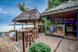 An outdoor seating area with wooden tables and chairs, a thatched-roof structure, and a view of the sea.