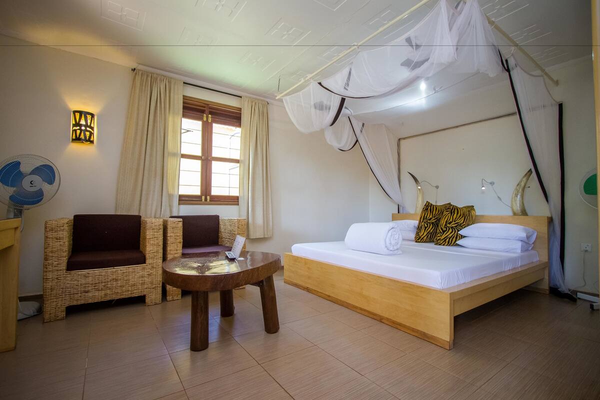 A room with a bed, two wicker chairs, a small round table, a wall-mounted fan, and a window with curtains.