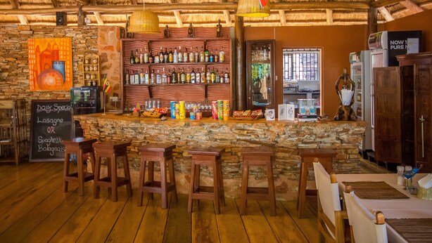 A rustic bar area with wooden stools, a stone wall, and a variety of bottles and snacks on shelves. - 2 Friends Guest House (Jinja)
