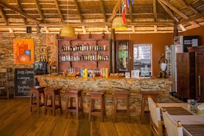 A rustic bar area with wooden stools, a stone wall, and a variety of bottles and snacks on shelves. - 2 Friends Guest House (Jinja)
