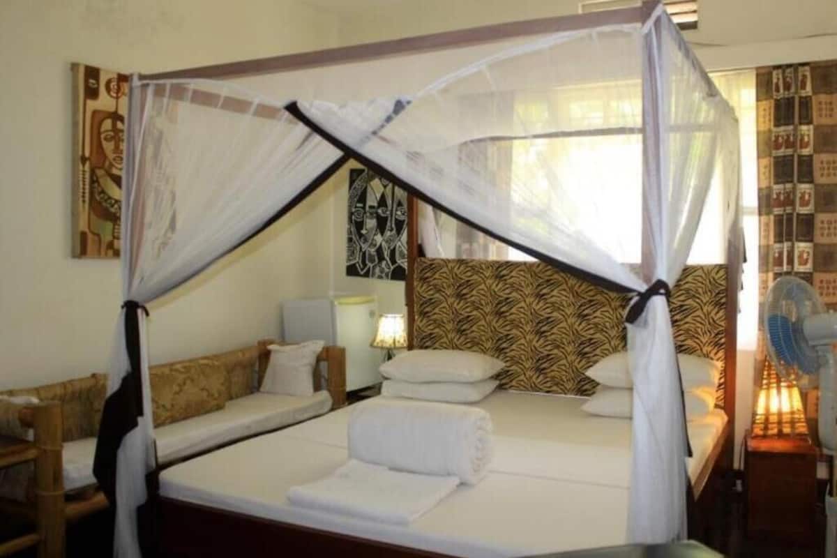 A room with a bed under a mosquito net, a sofa, a nightstand with a lamp, and a wall with patterned wallpaper.