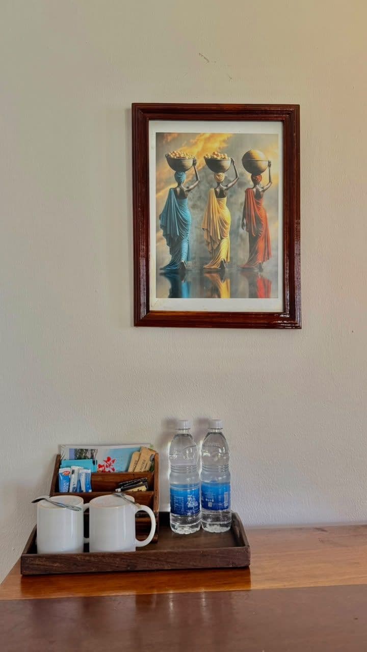 Une œuvre d’art encadrée représentant trois personnages sur un mur, un plateau avec des tasses et des bouteilles d’eau, et une table en bois.