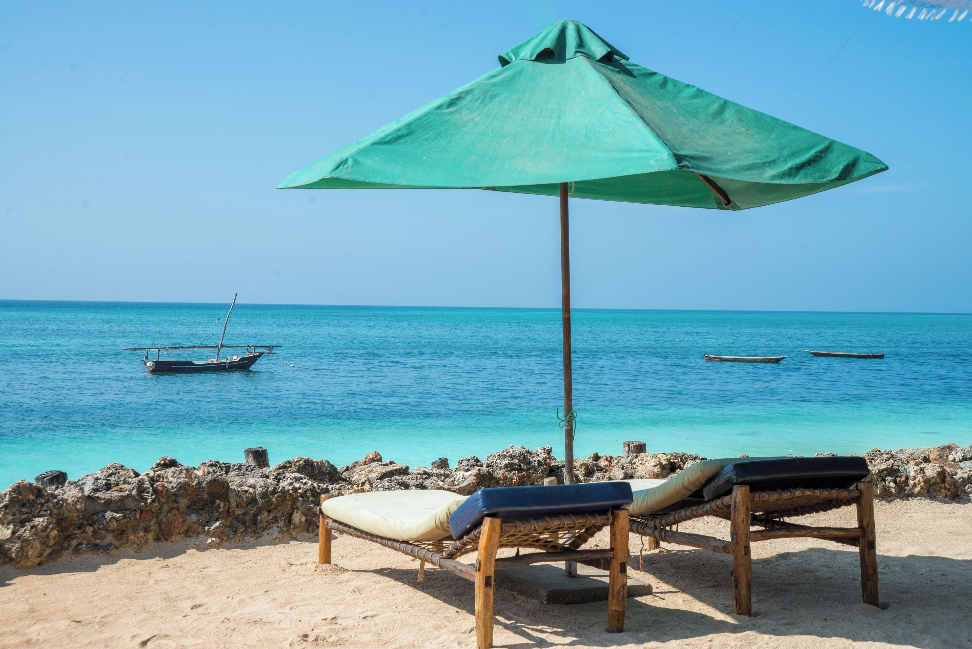 A beach with two wooden lounge chairs under a green umbrella, a rocky shoreline, and a calm sea with boats.