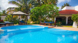 A swimming pool with lounge chairs and umbrellas, surrounded by tropical plants and a building with a red roof. - Flame Tree Cottages (Nungwi)