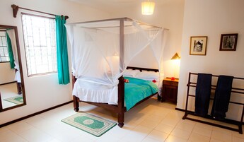 A four-poster bed with a mosquito net, a bedside table with a lamp, and a wooden chair with a towel.