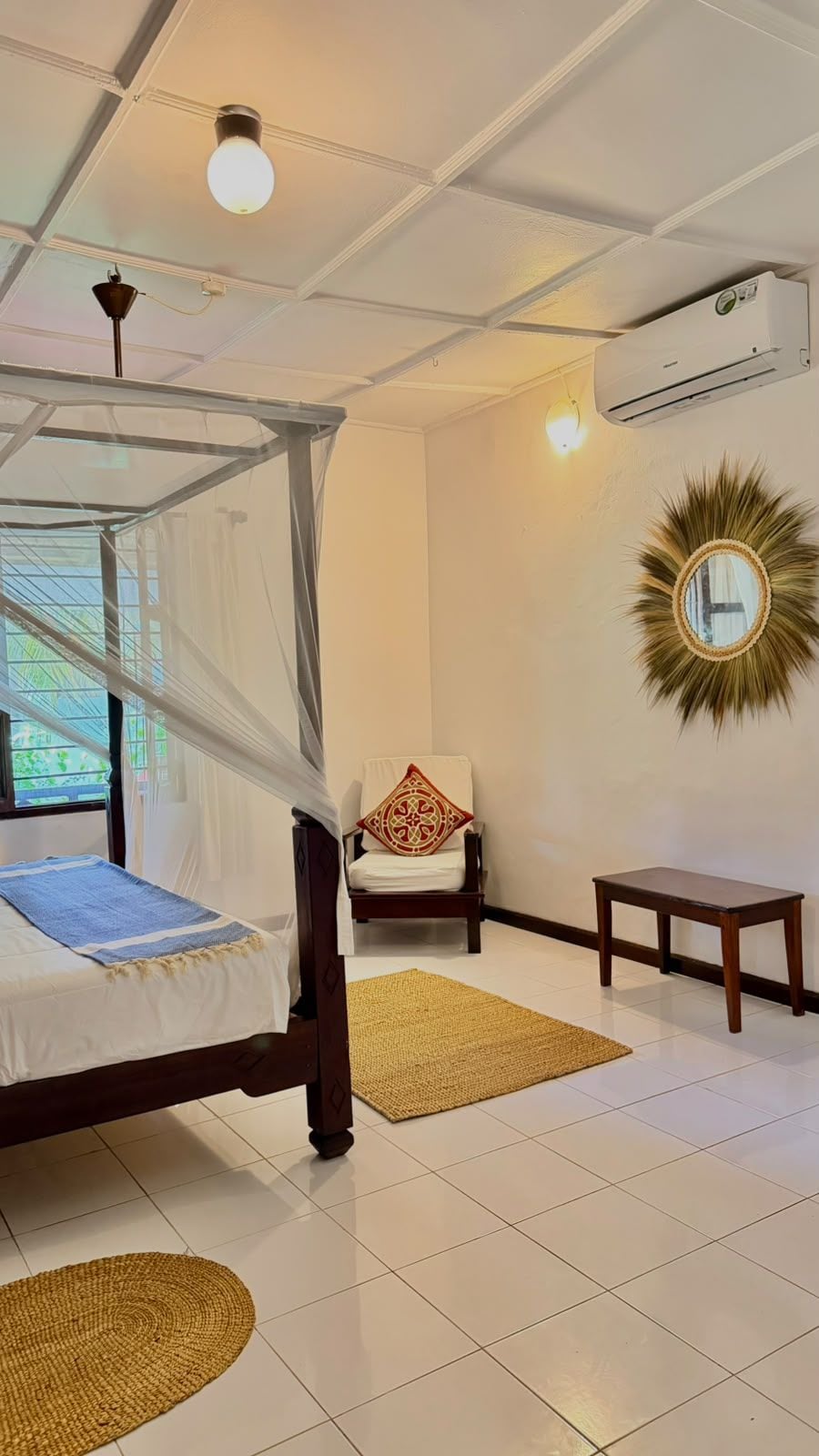 A bedroom with a bed, a chair, a small table, a decorative mirror, and an air conditioning unit.
