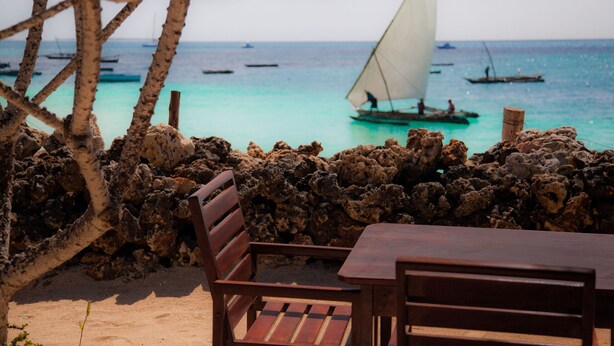 A beach view with a wooden table and chairs, rocky shoreline, and sailboats on the turquoise sea. - Flame Tree Cottages (Nungwi)