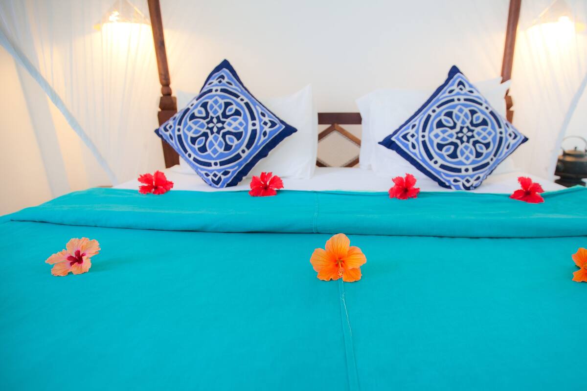 Una cama con una colcha turquesa, dos almohadas decorativas con diseños intrincados y varias flores de hibisco.