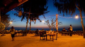 An outdoor dining area with tables and chairs, string lights, and a view of the ocean at dusk. - Flame Tree Cottages (Nungwi)