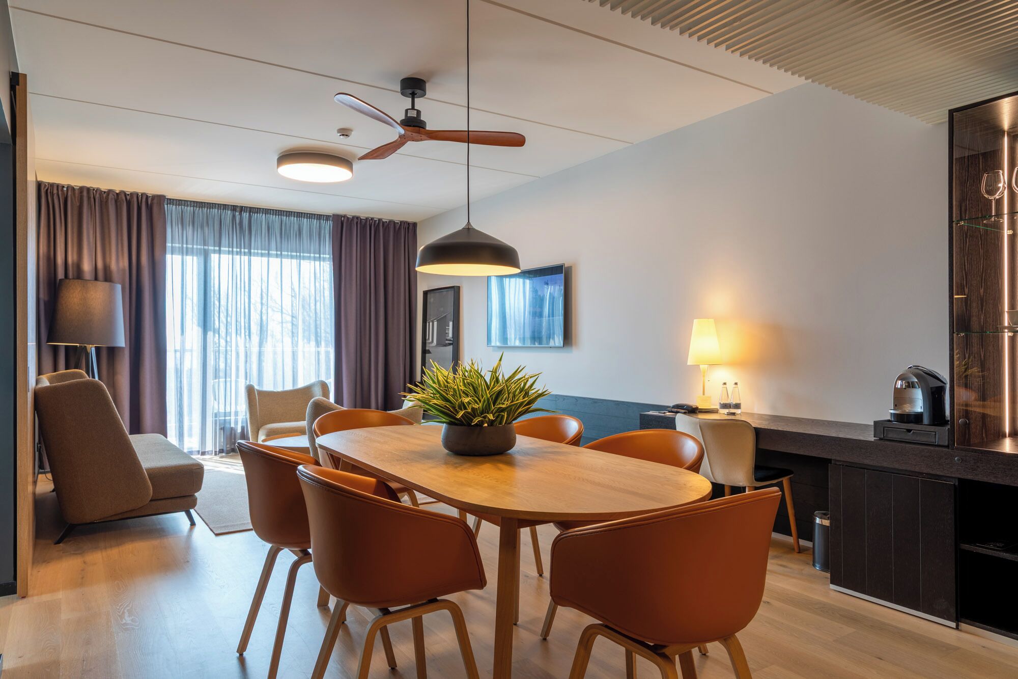 Quarto de hotel moderno com área de jantar, ventilador de teto, planta em vaso e televisão fixada na parede.