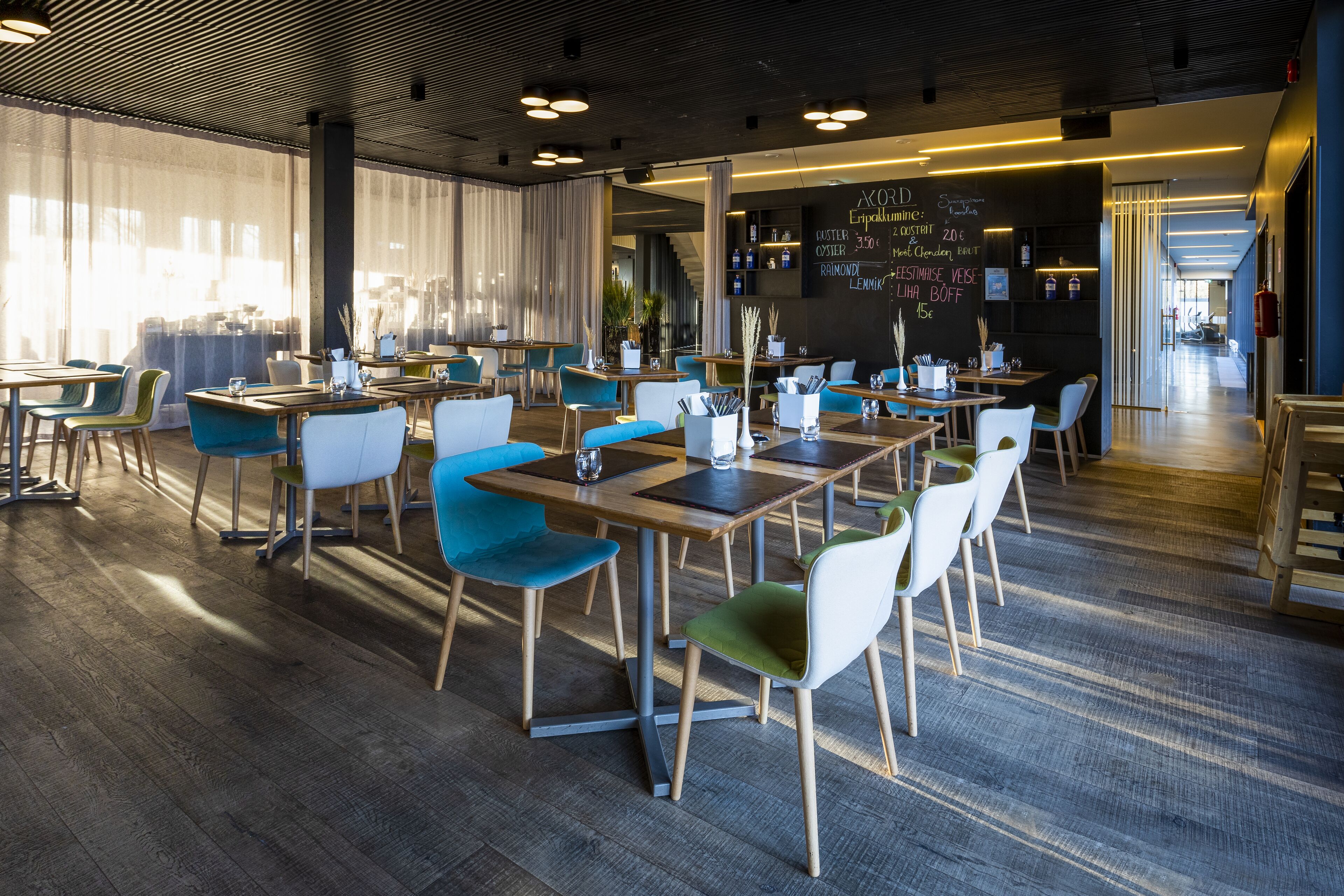 En moderne restaurant med træborde og stole, en tavle med menu og store vinduer, der lukker naturligt lys ind.