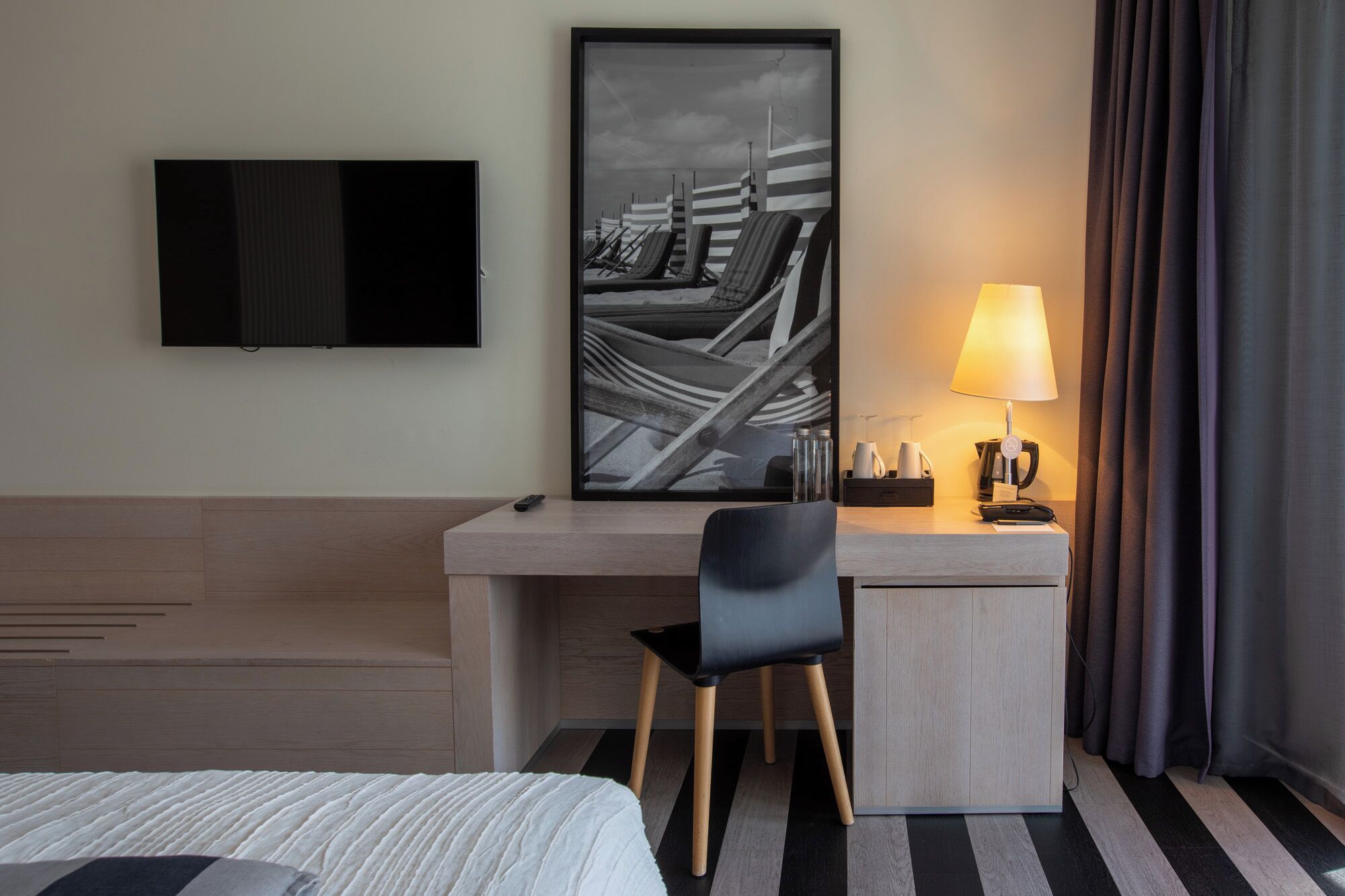 A hotel room with a bed, a desk, a chair, a television, a framed artwork, a lamp, and a window with curtains.