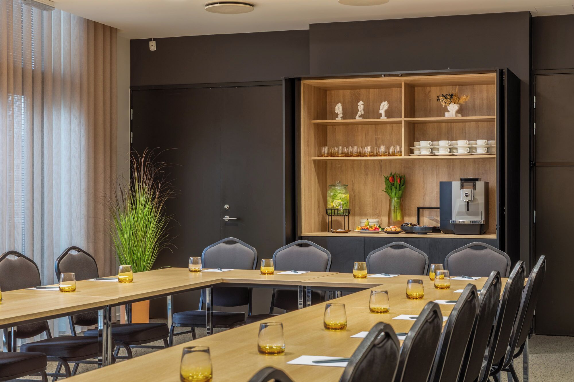 A conference room with a long table, chairs, a coffee station, and a decorative shelf with items.