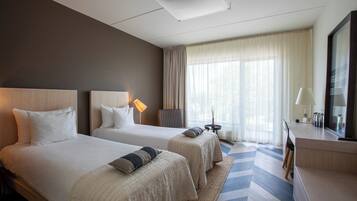 Une chambre d’hôtel avec deux lits, un bureau, une chaise, une lampe et une grande fenêtre avec des rideaux.