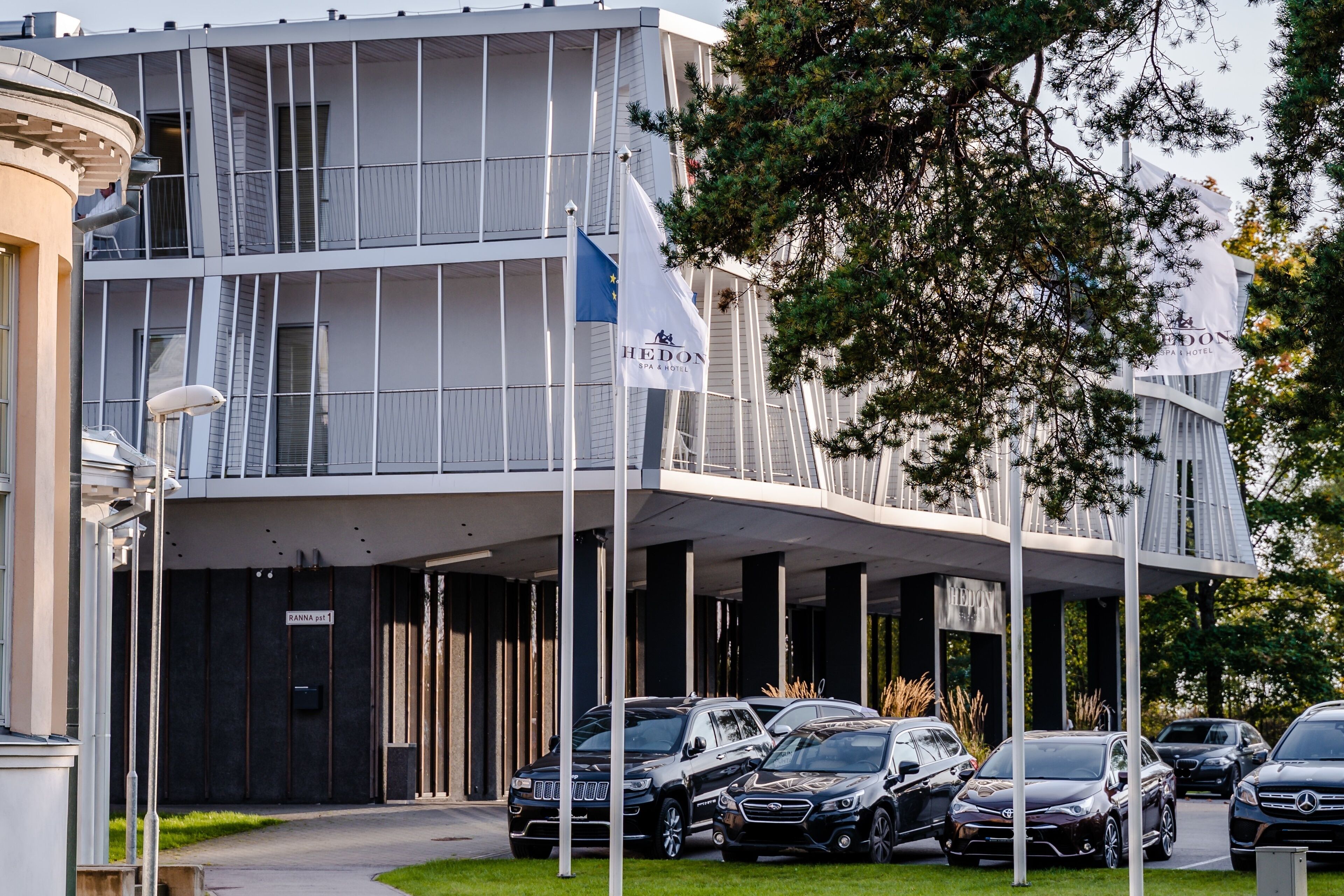A modern hotel with a covered entrance, multiple cars parked in front, and a flag indicating European location.