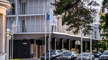 A modern hotel with a covered entrance, multiple cars parked in front, and a flag indicating European location.