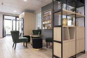 Un interior moderno con un mostrador de recepción, sillas tapizadas en verde, una pequeña mesa redonda y una estantería de madera con objetos decorativos.