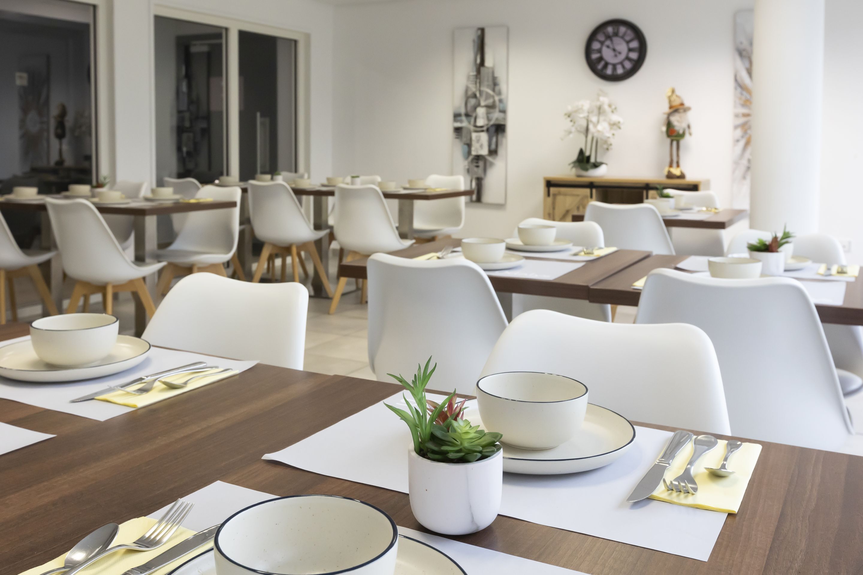 Een eetruimte met meerdere tafels, witte stoelen en een klok aan de muur.