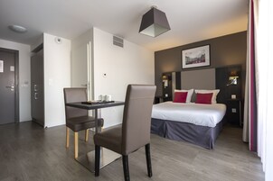 A modern hotel room with a bed, desk, chair, and a dining area.