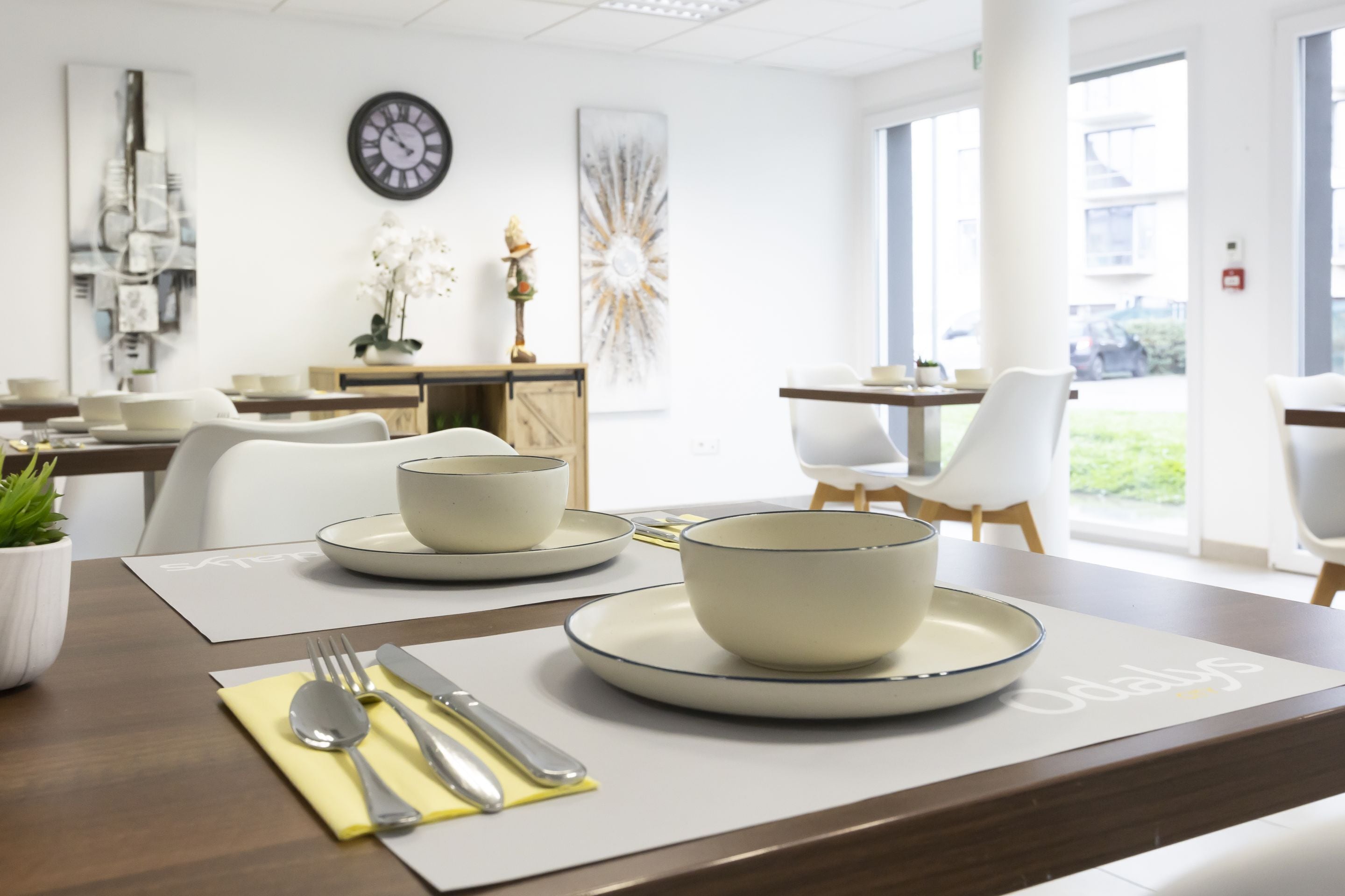 A modern dining area with a wooden table set for two, featuring white plates and bowls, and a clock on the wall.