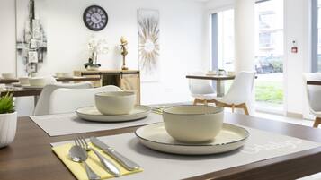 A modern dining area with a wooden table set for two, featuring white plates and bowls, and a clock on the wall.