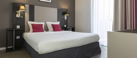 Una habitación de hotel moderna con una cama grande, dos mesitas de noche con lámparas, un cuadro enmarcado en la pared y una ventana con cortinas.