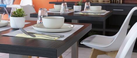 A dining area with tables set for two, white chairs, a flat-screen TV mounted on the wall, and a small potted plant on the table.