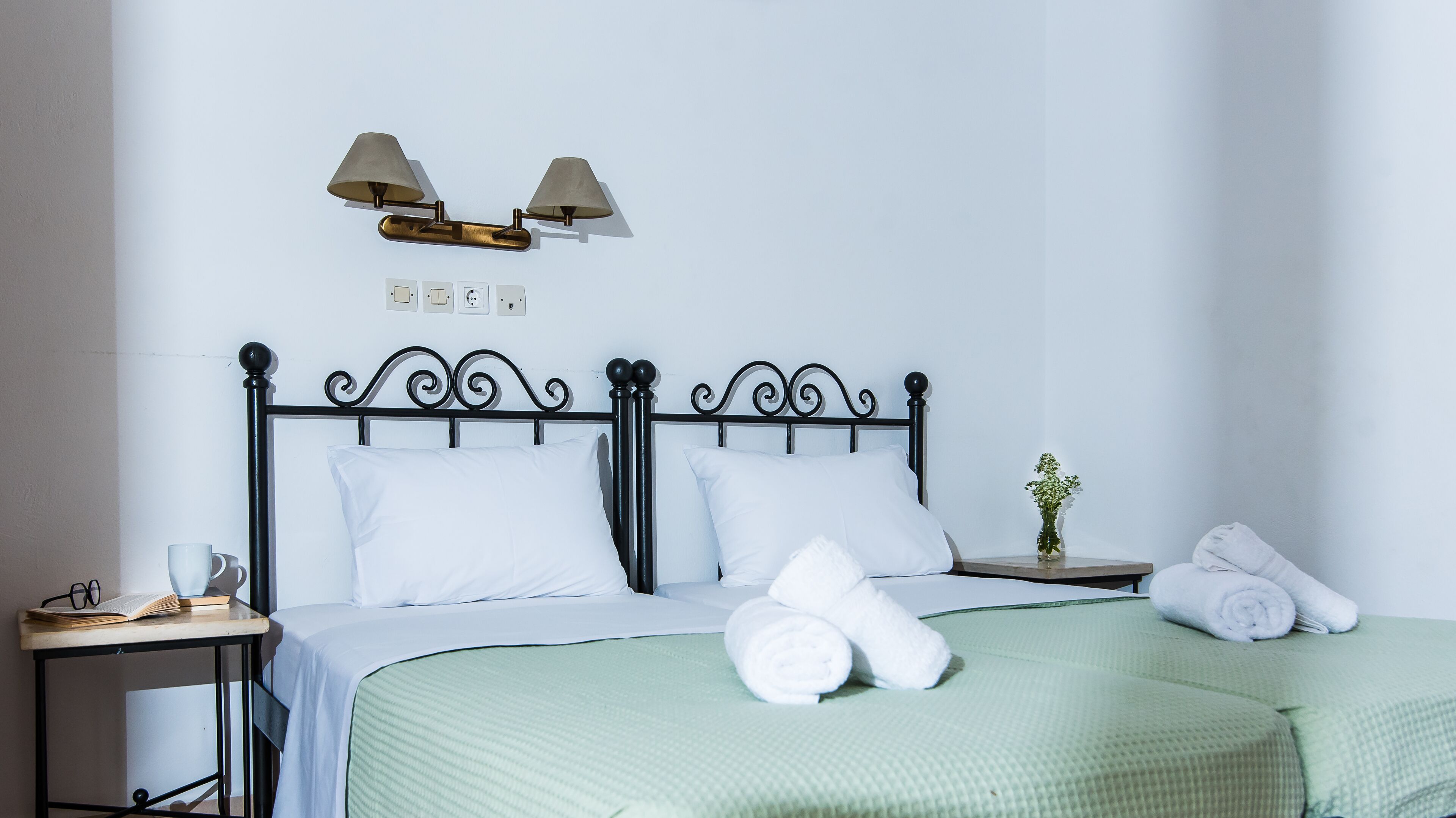 A neatly made bed with white linens, a green textured bedspread, and two rolled towels on the side. There is a small bedside table with a book, glasses, and a cup. A wall-mounted lamp with two shades is above the bed.