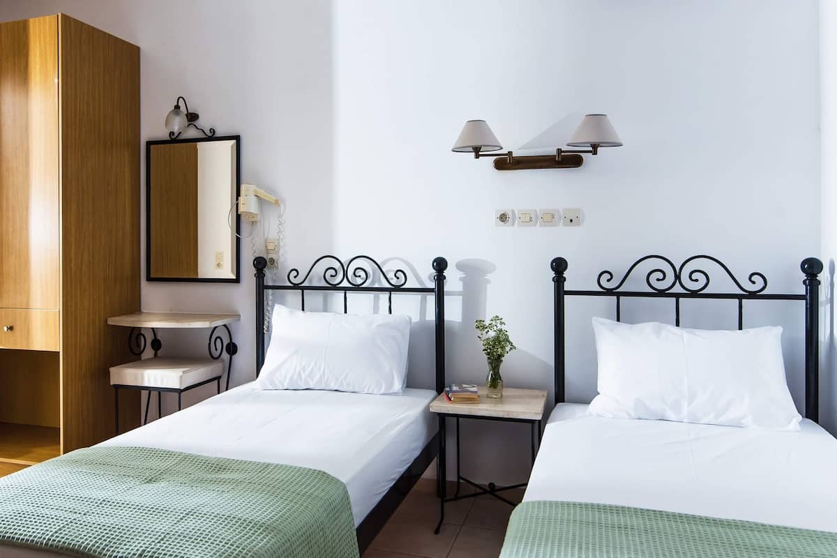 A hotel room with two single beds, a wooden wardrobe, a small bedside table with a lamp, and a mirror on the wall.