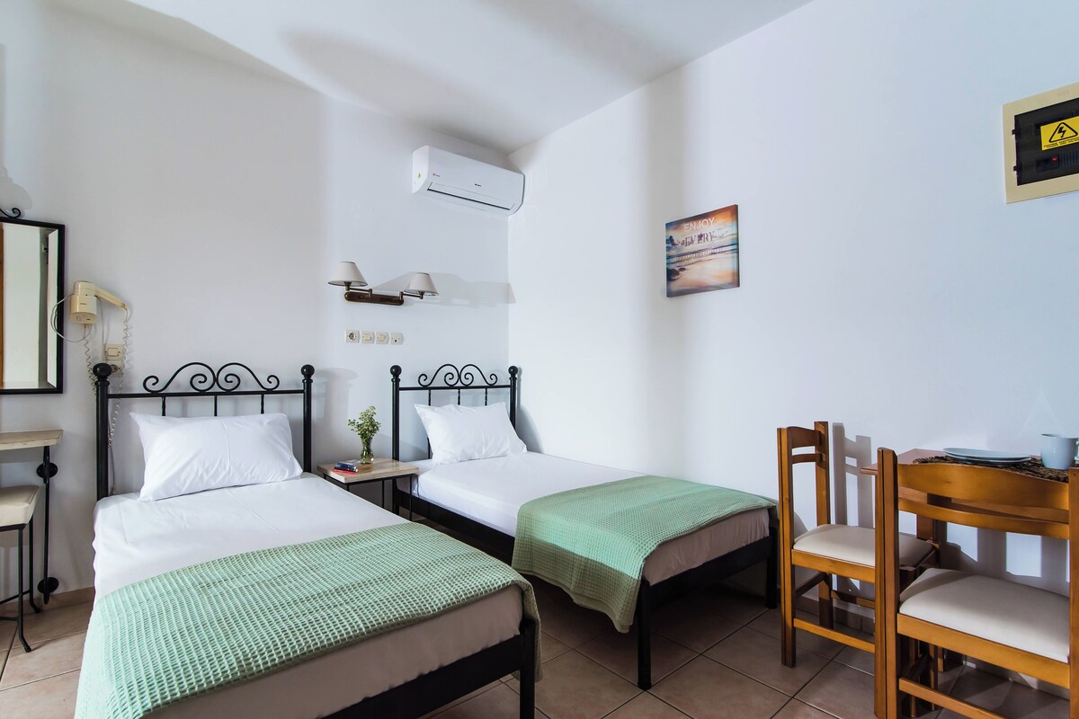 A room with two single beds, a desk with a chair, a small table with a lamp, an air conditioning unit, and a wall-mounted electrical outlet.