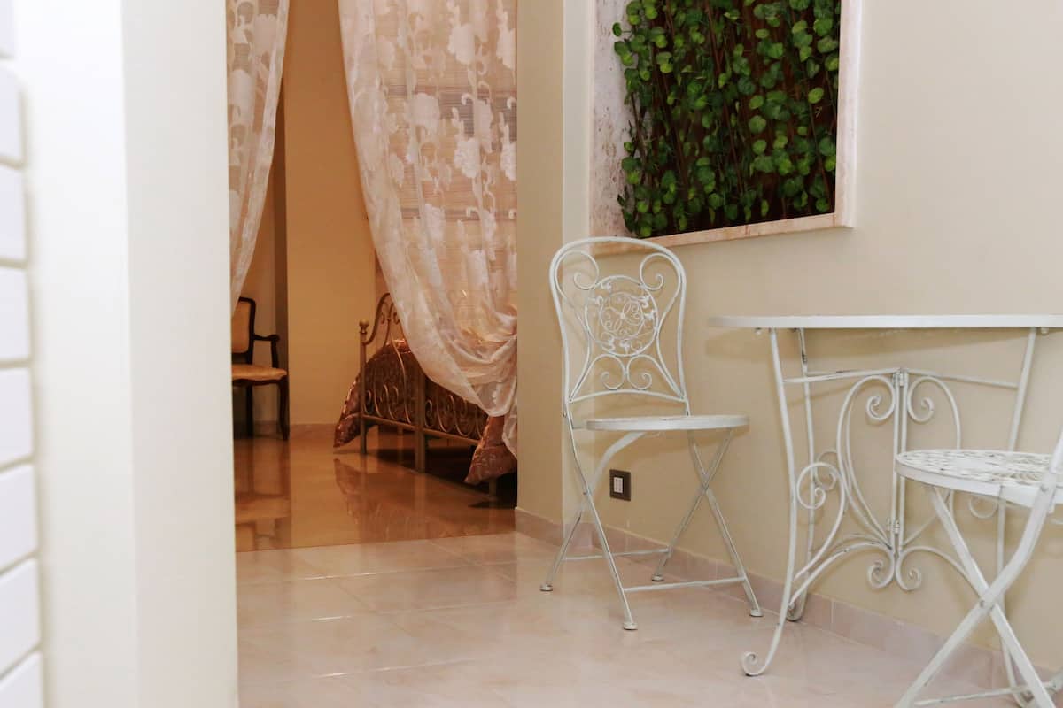 Une pièce avec un sol carrelé de carreaux blancs, une chaise en métal blanche, une petite table blanche et un lit surmonté d’un baldaquin blanc.