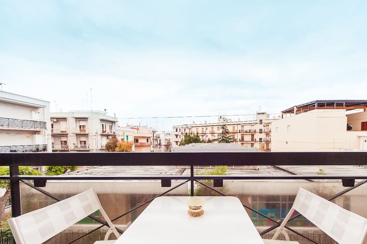 Un balcon avec une table et des chaises, donnant sur un quartier résidentiel avec des immeubles et un ciel dégagé.