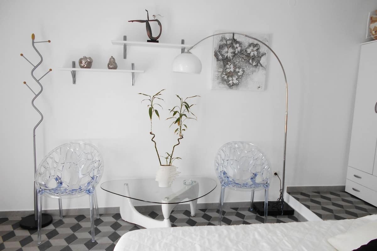 A modern room with a glass table, two chairs, a bed, a floor lamp, and wall-mounted shelves with decorative items.