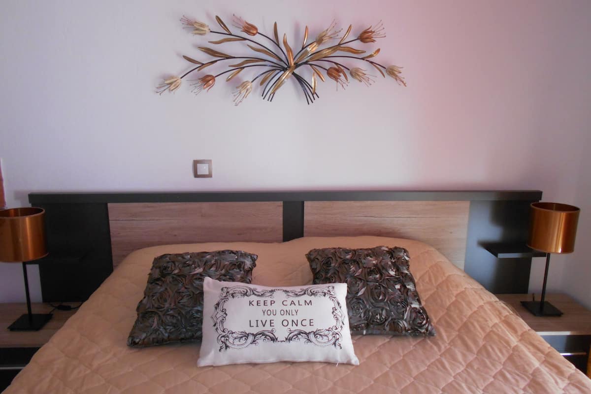 A bed with a beige quilted bedspread, two decorative pillows with a floral pattern, and a wall art piece above the bed.
